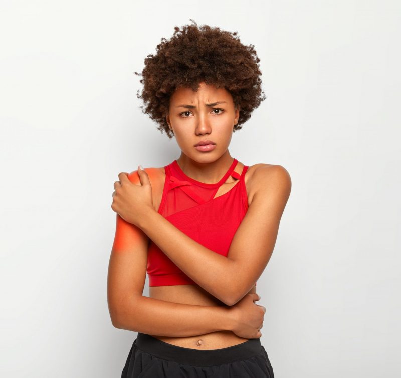Photo of sorrowful dark skinned woman cries from terrible pain, touches red blades, dressed in casual wear, shows bare shoulders, isolated over white background. Office syndrom and stiffness